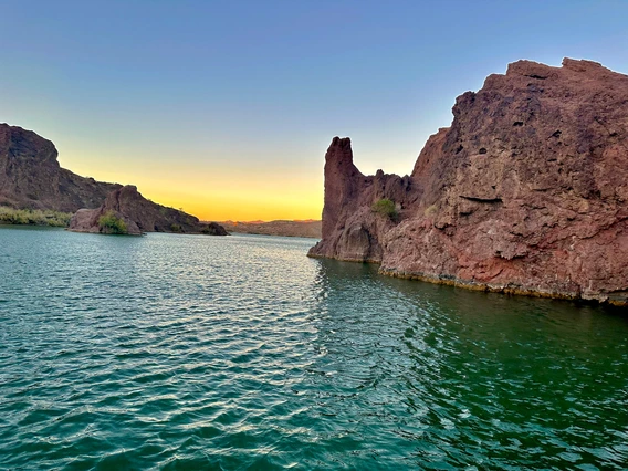 photo of sunset on lake havasu. Water looks very green, and there are red rock formations bordering it. The sunset is yellow with some orange banding