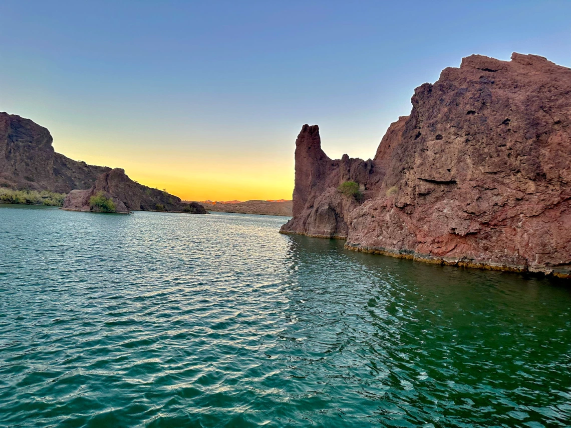 photo of sunset on lake havasu. Water looks very green, and there are red rock formations bordering it. The sunset is yellow with some orange banding