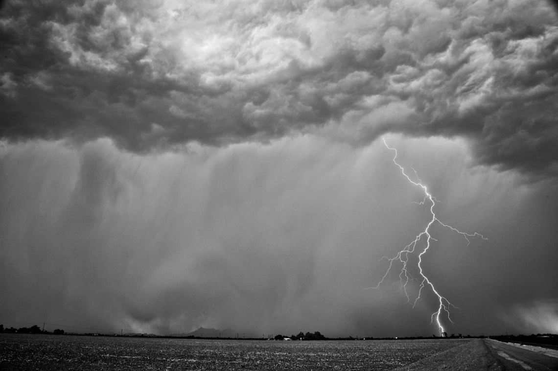 black and white photo showing an agricultural landscape with showing rain and a single lightning bolt